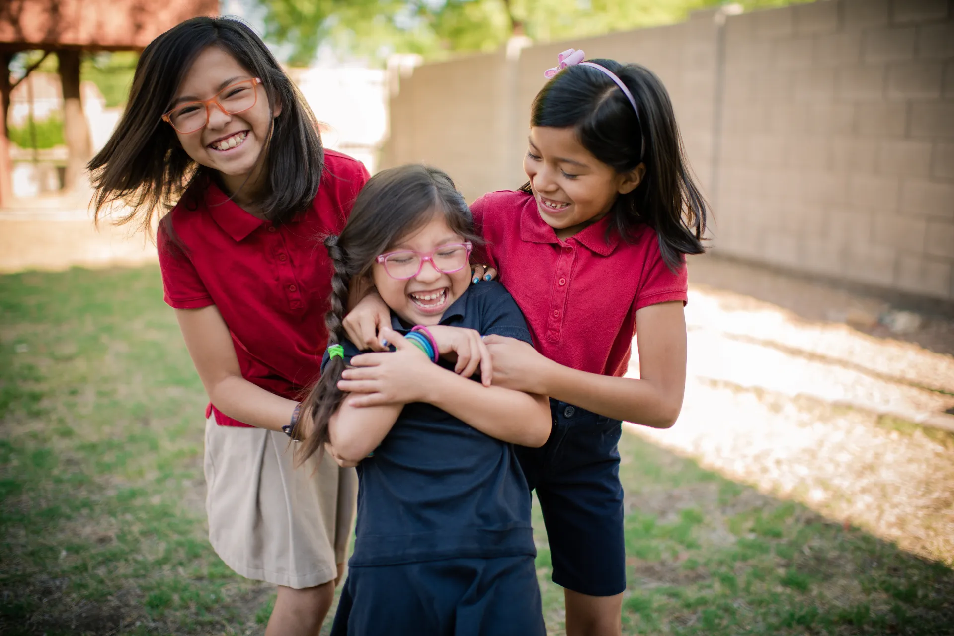 Tres niñas riendo y abrazándose en un patio, comunidad de familias